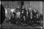 Vecpiebalgas ev.-lut.baznīcas mācītājs Augusts Trucis ar jauniešiem. No kreisās 1.rindā 4. - Alberts Kanders, 3.rindā 4.-Agnese Kandere no "Mierupītes" mājām. Fonā koka ēka ar pavērtām durvīm. Visi jaunekļi uzvalkos. Foto - Gustavs Žakerts